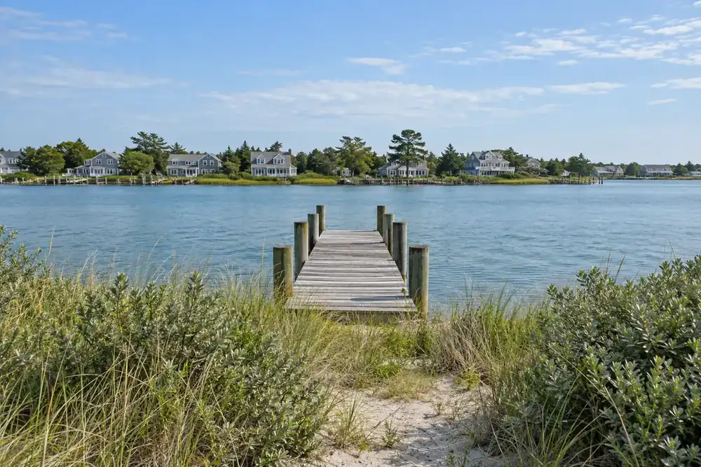 Bayberry shrubs and dune grass beside water, a wooden dock and distant shingled houses